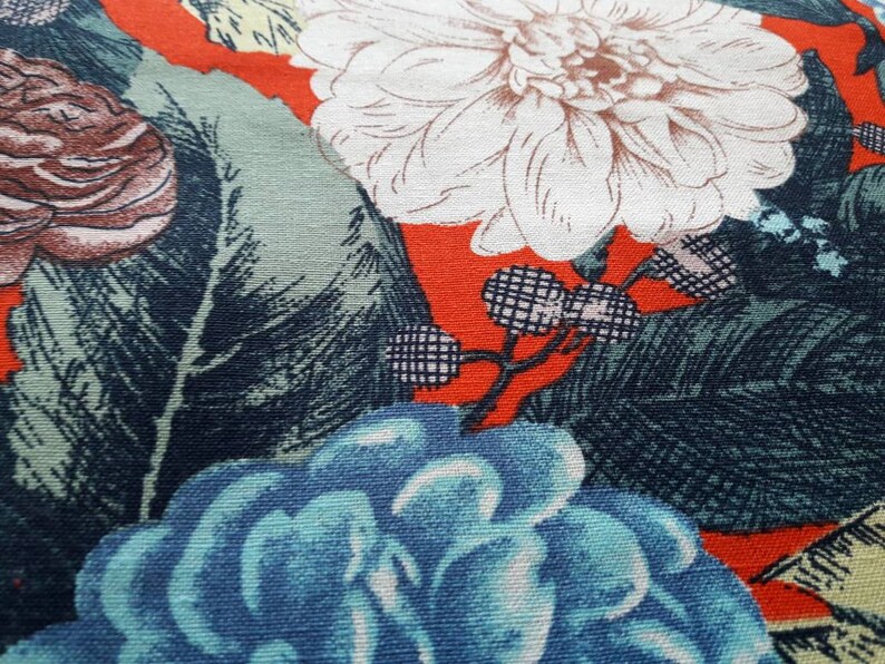 May include: A close-up of a fabric with a floral pattern. The fabric features a large white flower with a red center, a blue flower, and green leaves on a red background.