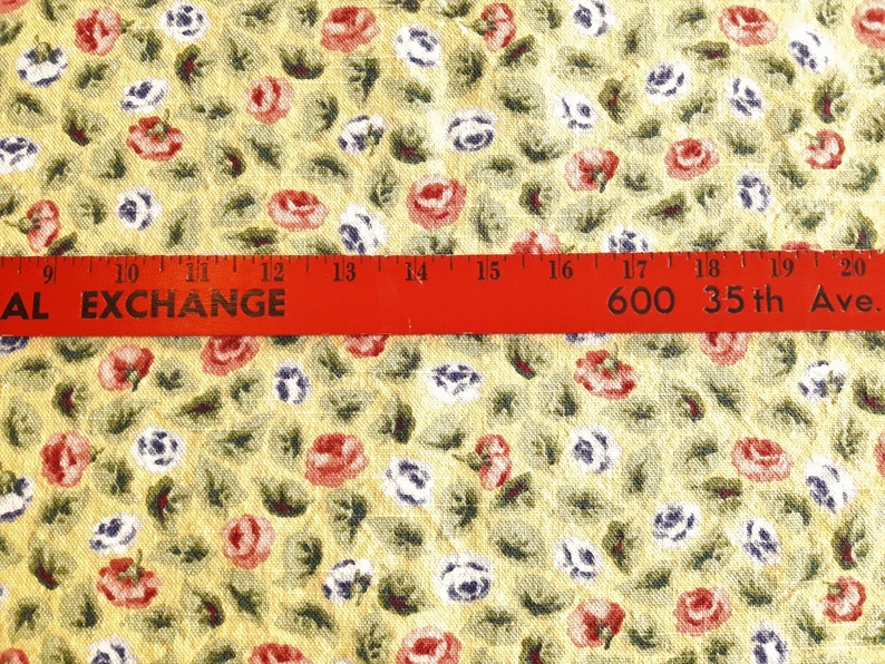 May include: Yellow fabric with a repeating pattern of small red, white, and blue flowers. A red ruler with the text "AL EXCHANGE 600 35th Ave." is placed across the fabric.