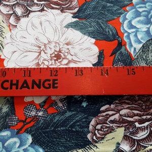May include: Red fabric with a floral print featuring white, blue, and brown flowers. A red ruler with the word "EXCHANGE" printed on it is placed across the fabric. The ruler measures 10 to 16 inches.