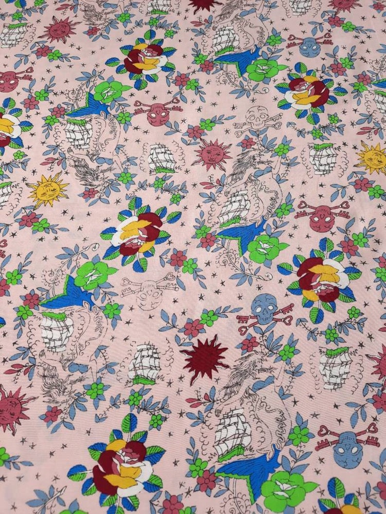 May include: Pink fabric with a repeating pattern of traditional tattoo imagery. The design includes roses, swallows, ships, skulls, keys, suns, and stars in red, yellow, green, blue, and black.