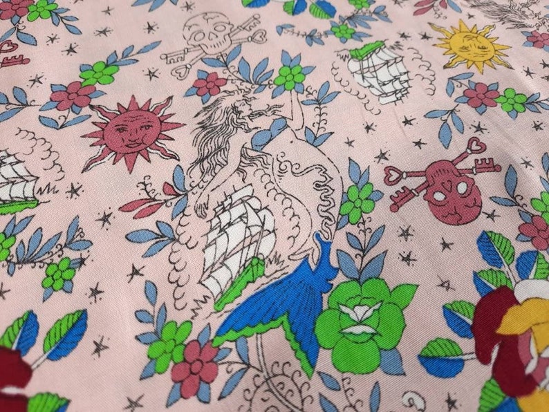 May include: A light pink fabric with a repeating pattern of nautical and pirate-themed illustrations. The design includes a mermaid, ships, suns, skulls with crossed keys, and floral elements in various colors.