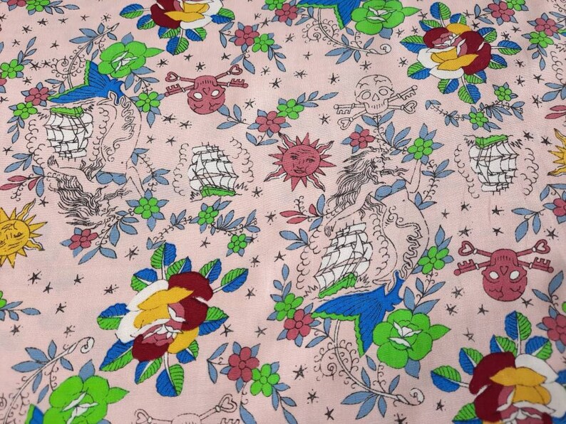 May include: Pink fabric with a repeating pattern of nautical and floral elements. The design includes sailing ships, roses, skulls with crossed bones, suns, and mermaids, in shades of red, yellow, blue, and green.