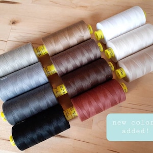 May include: A collection of spools of thread in various shades of gray, brown, and beige. The spools are arranged in a semi-circle on a wooden surface. The text "new colors added!" is written in a blue box on the right side of the image.