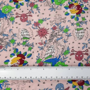 May include: Pink fabric with a repeating pattern of nautical and floral elements. The design includes ships, roses, skulls with keys, suns, and mermaids. The color palette features pink, blue, green, yellow, and red. A ruler is placed on the fabric, showing inches.
