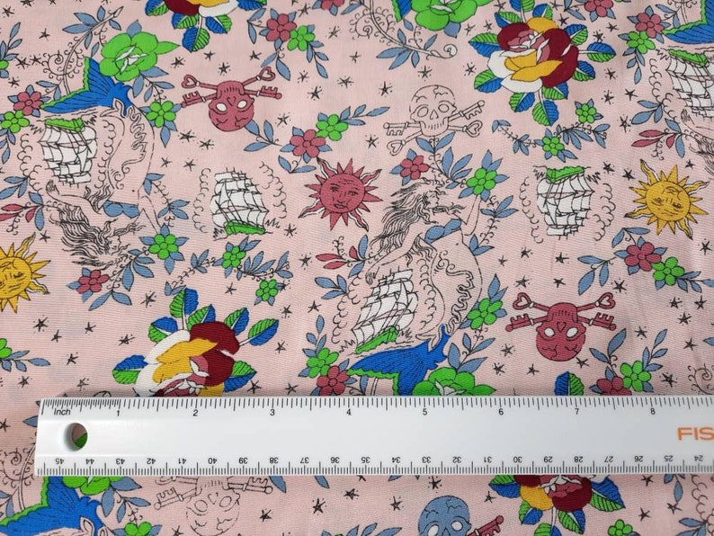 May include: Pink fabric with a repeating pattern of nautical and floral elements. The design includes ships, roses, skulls with keys, suns, and mermaids. The color palette features pink, blue, green, yellow, and red. A ruler is placed on the fabric, showing inches.