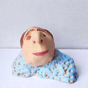 Peut inclure: Une figurine en argile sculptée à la main représentant une tête aux cheveux bruns, au nez proéminent et aux lèvres roses, fixée sur une base bleu clair texturée avec de petites indentations beige. La figure regarde vers le haut.