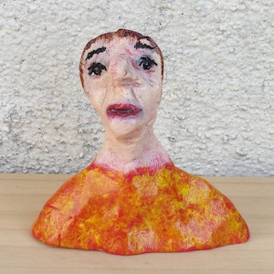 May include: A handmade sculpture of a human bust. The face has a distressed expression with dark eyes and red lips. The figure wears an orange and yellow top. The sculpture is on a wooden surface.