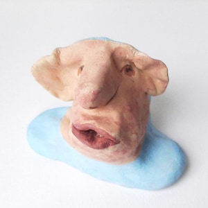 Peut inclure: Une figurine en argile sculptée à la main présentant un visage pâle et texturé avec de grandes oreilles, un nez proéminent et des lèvres pleines rose foncé. La tête est fixée sur une base lisse bleu clair, formant un buste de personnage détaillé.