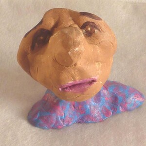 May include: A handmade clay sculpture of a face with brown skin, dark brown eyebrows, and pink lips. The sculpture has a blue and pink patterned collar. The face has a prominent nose and is slightly tilted.