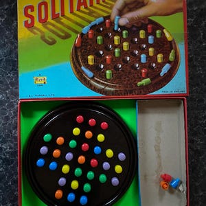 Pode incluir: Um jogo de paciência Merit na sua caixa original. O tabuleiro de jogo é feito de plástico preto com um design circular. A caixa apresenta uma imagem colorida do tabuleiro de jogo com o texto "Merit Solitaire" em letras vermelhas.