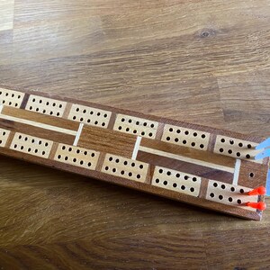Könnte beinhalten: Ein hölzernes Cribbage-Brett mit rechteckiger Form und einer Vielzahl von Bohrlöchern. Das Brett hat eine Mischung aus hellen und dunklen Holztönen, mit blauen und roten Stiften am Ende.