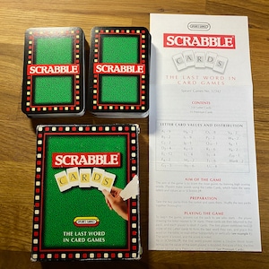 Pode incluir: Uma caixa de jogo de cartas Scrabble com um fundo verde e o texto "Scrabble Cards The Last Word in Card Games". A caixa é cercada por duas pilhas de cartas com o texto "Scrabble" nelas.