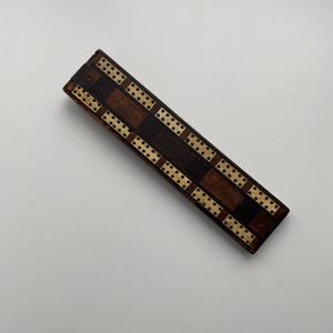 Könnte beinhalten: Ein rechteckiges Cribbage-Brett aus Holz mit Schachbrettmuster. Das Brett besteht aus dunkelbraunem und hellbraunem Holz mit Reihen kleiner Löcher für die Stifte. Das Brett ist etwa 30 cm lang.