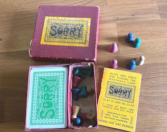 Könnte beinhalten: Vintage "Sorry!" Brettspiel-Set. Die bordeauxfarbene Box hat ein goldenes Etikett. Das Spiel enthält ein Kartenspiel mit grün-weißen Designs, mehrfarbige Spielfiguren und ein gelbes Anleitungsheft.