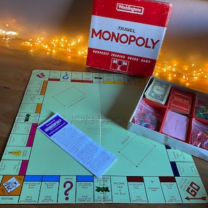 Könnte beinhalten: Ein Vintage Travel Monopoly Brettspiel-Set. Die rot-weiße Box ist geöffnet und zeigt Spielsteine, Karten und Würfel. Das Spielbrett ist hellgrün mit Spielfeldern und Anweisungen. Ein klassisches Familienspiel.
