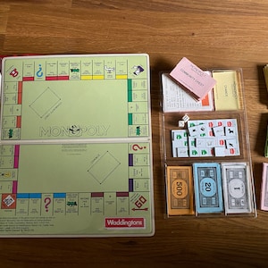 Könnte beinhalten: Ein Vintage-Monopoly-Brettspiel mit einem grünen Spielbrett, roten und gelben Eigentumsbriefen und Geldscheinen in Stückelungen von 1, 5, 10, 20 und 500. Das Spiel enthält Chance- und Gemeinschaftskarten, Würfel und Eigentumssteine.