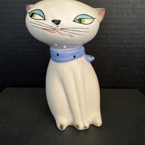 Puede incluir: Figura de gato de cerámica blanca con bufanda azul. El gato tiene ojos verdes, bigotes negros y boca rosa. La figura es un diseño estilizado de mediados de siglo.