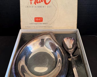 May include: A silver serving set in a white box. The set includes a small, teardrop-shaped dish and a spoon. The box has the words "Flair Entertainment Set" and "1847 Rogers Bros." printed on it.