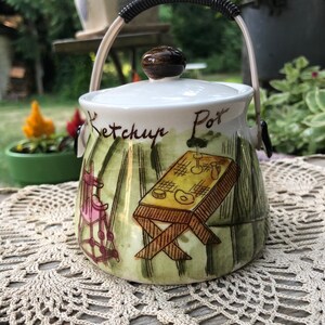 May include: A white ceramic ketchup pot with a black handle and a green, yellow, and brown painted design. The pot has the words "Ketchup Pot" written on it.