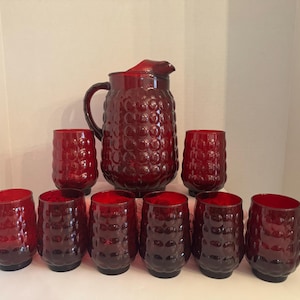 May include: A vintage red glass drinkware set, including a pitcher and eight matching tumblers. The pitcher has a handle and a spout. The glasses and pitcher have a textured, bubble-like pattern. The set is ideal for serving beverages.