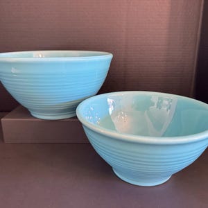 May include: Two turquoise ceramic bowls. The bowls have a rounded shape with horizontal ridged detailing. The larger bowl sits on a small, brown rectangular base. The bowls are empty and have a glossy finish.