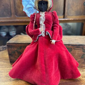 May include: A vintage doll wearing a red velvet dress with a white lace trim and a blue and white umbrella. The doll has a black face and is made of fabric.