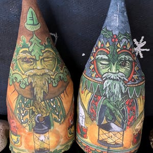 Puede incluir: Dos adornos de tela en forma de cono con ilustraciones impresas de figuras barbudas. La figura de la izquierda es marrón y verde con un motivo de bellota. La figura de la derecha es azul y verde con hojas de acebo y bayas, ambas sostienen linternas.