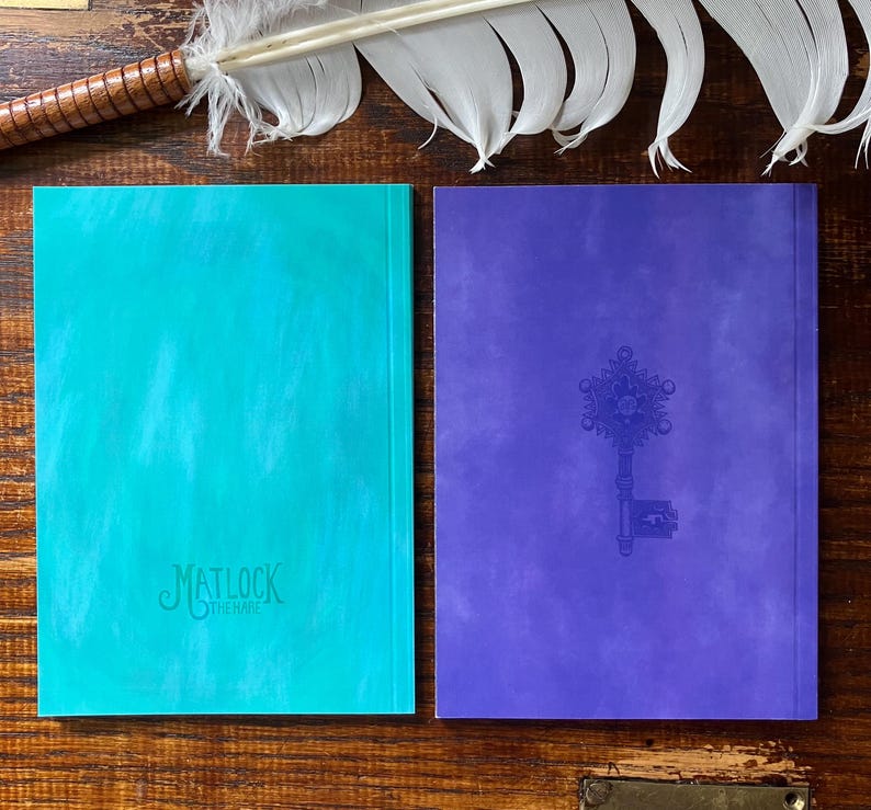 May include: Two hardcover journals. One is teal with the text "Matlock the Hare" embossed on the cover. The other is purple with an embossed key design. A feather quill rests above the journals.