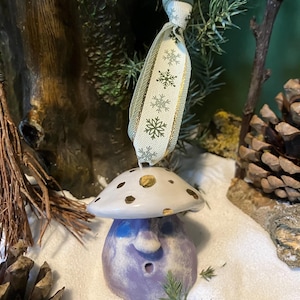 Puede incluir: Adorno de hongo de cerámica caprichoso con una tapa blanca con lunares dorados y una base lavanda. El adorno tiene una cara y está suspendido por una cinta con acentos de copo de nieve. El fondo incluye piñas y vegetación.