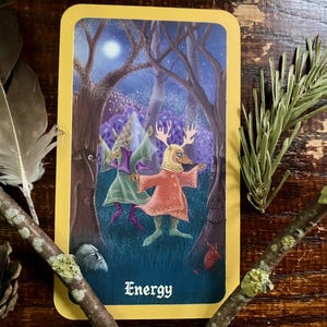 May include: A tarot card with a yellow border features an illustration of a figure with antlers and a red dress in a forest setting. The card is labeled "Energy" in a stylized font. The image is surrounded by natural elements like feathers and twigs.