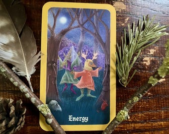May include: A tarot card with a yellow border features an illustration of a figure with antlers and a red dress in a forest setting. The card is labeled "Energy" in a stylized font. The image is surrounded by natural elements like feathers and twigs.