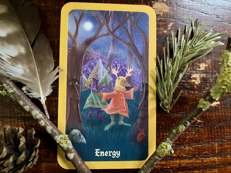 May include: A tarot card with a yellow border features an illustration of a figure with antlers and a red dress in a forest setting. The card is labeled "Energy" in a stylized font. The image is surrounded by natural elements like feathers and twigs.