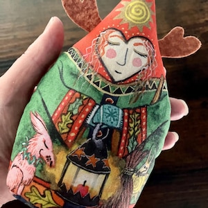 Könnte beinhalten: Ein weiches Stoffornament in Form einer spitzen Mütze mit braunem Filzgeweih. Der Druck zeigt eine Person mit roten Haaren, einem grünen Umhang, einer Laterne und einem kleinen rosa Tier. Das Design enthält eine gelbe Sonne und Herbstblätter.