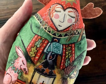 May include: A soft, stuffed fabric ornament shaped like a pointed hat with brown felt antlers. The print features a person with red hair, a green cloak, a lantern, and a small pink animal. The design includes a yellow sun and autumn leaves.