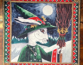May include: A square fabric wall hanging featuring a white rabbit dressed as a witch in a green hat and robe, holding a broomstick. The scene includes a full moon, snowy landscape, and a decorative border with red and green geometric patterns.