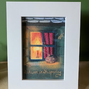 May include: A framed artwork featuring a cozy autumn scene. The image depicts a window with a lit candle, red curtains, and a decorative pumpkin. The text "Autumn is whispering" is visible. The frame is white.