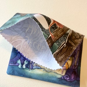May include: A small, blue fabric pouch with a whimsical illustration of a witch and her cauldron. The pouch is folded in half, revealing a white fabric lining with the word "Magic" written in cursive.