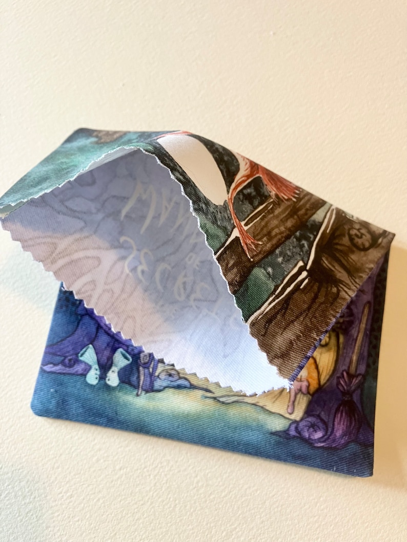 May include: A small, blue fabric pouch with a whimsical illustration of a witch and her cauldron. The pouch is folded in half, revealing a white fabric lining with the word "Magic" written in cursive.