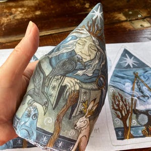 Puede incluir: Un cono de tela con una ilustración impresa de una figura anciana con cabello blanco, una corona y una capa oscura, acompañada por un cuervo negro, un conejo y una criatura azul. El diseño incluye una estrella, patrones y un borde.
