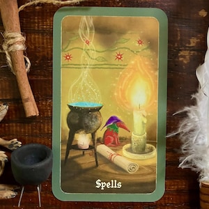 May include: A tarot card with a green border features a cauldron with blue liquid, a lit candle, and a scroll. A small figure in a hat and a fox figurine are also present. The word "Spells" is at the bottom of the card.