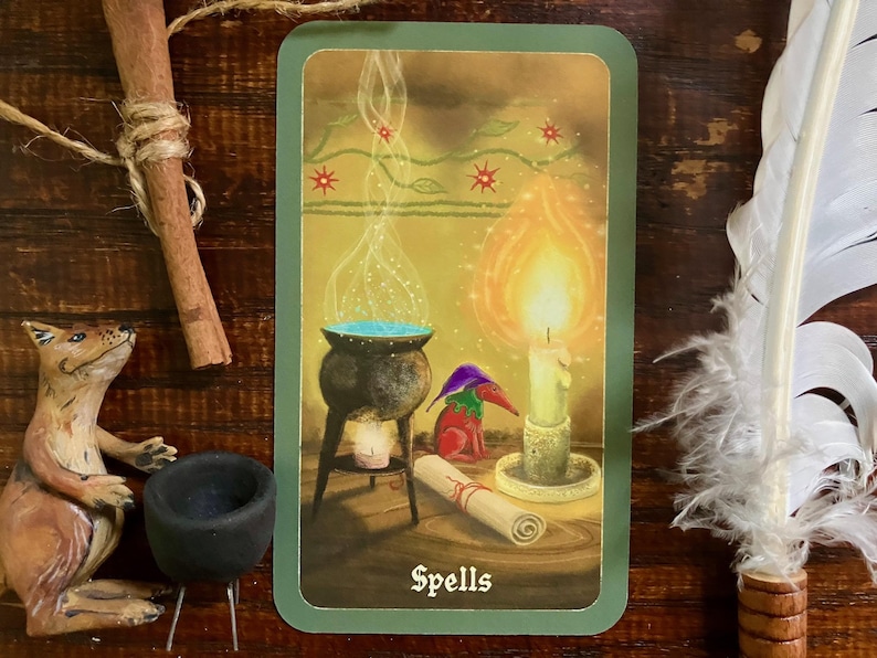 May include: A tarot card with a green border features a cauldron with blue liquid, a lit candle, and a scroll. A small figure in a hat and a fox figurine are also present. The word "Spells" is at the bottom of the card.
