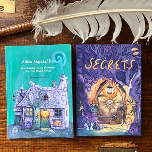 Peut inclure: Deux livres illustrés. L'un a une couverture turquoise avec le titre "A Most Majickal Year". L'autre livre est intitulé "SECRETS" avec une couverture violette. Une plume blanche est en arrière-plan. Les livres présentent des illustrations fantastiques.