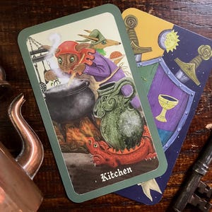 May include: Two tarot cards with illustrations of fantasy creatures and symbols. One card depicts a creature cooking in a cauldron, labeled "Kitchen." The other shows a shield, swords, and a chalice. A copper teapot and key are also visible.