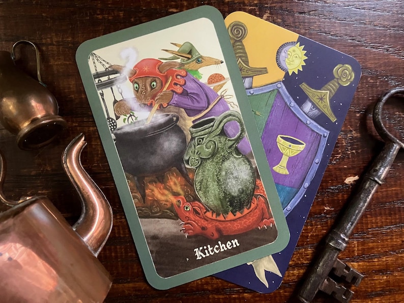 May include: Two tarot cards with illustrations of fantasy creatures and symbols. One card depicts a creature cooking in a cauldron, labeled "Kitchen." The other shows a shield, swords, and a chalice. A copper teapot and key are also visible.