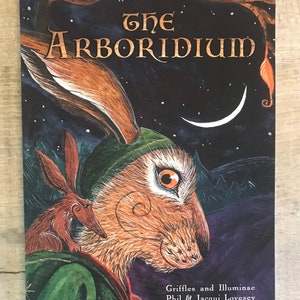 以下が含まれることがあります： 濃い青色の背景に三日月が描かれた本の表紙。タイトルの「The Arboridium」は金色の文字で書かれている。表紙には緑色の帽子をかぶった茶色のウサギと、その背中に乗った小さな茶色の生き物が描かれている。