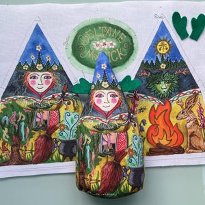Puede incluir: Muñeca de tela hecha a mano con un diseño caprichoso. Presenta una cara sonriente, un sombrero azul y una corona de hojas verdes. Ilustraciones de árboles, un caldero, una hoguera y un conejo rodean la muñeca. El texto "Beltane Magick" es visible.