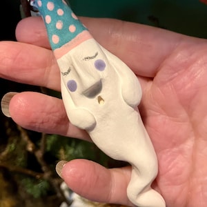 Puede incluir: Figura de fantasma de cerámica hecha a mano. El fantasma blanco tiene una cara sonriente, ojos morados y una pequeña boca dorada. Lleva un gorro de fiesta azul con lunares rosas y un borde rosa. La figura tiene un cuerpo curvado y alargado.