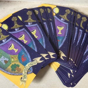 May include: A deck of illustrated cards with a yellow border. Each card features a shield design with a sword, chalice, and castle. The cards have a dark blue background with stars and a yellow sun and moon.
