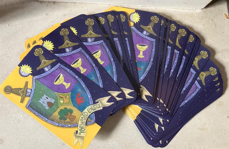 May include: A deck of illustrated cards with a yellow border. Each card features a shield design with a sword, chalice, and castle. The cards have a dark blue background with stars and a yellow sun and moon.