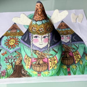Könnte beinhalten: Stoffpuppe mit vogelartigem Kopfschmuck und bemaltem Gesicht, die einen Korb hält. Die Puppe ist Teil einer größeren Illustration mit anderen Figuren und einer Landschaft mit Sonne, Bäumen und Tieren. Die Puppe hat weiße Filzflügel.